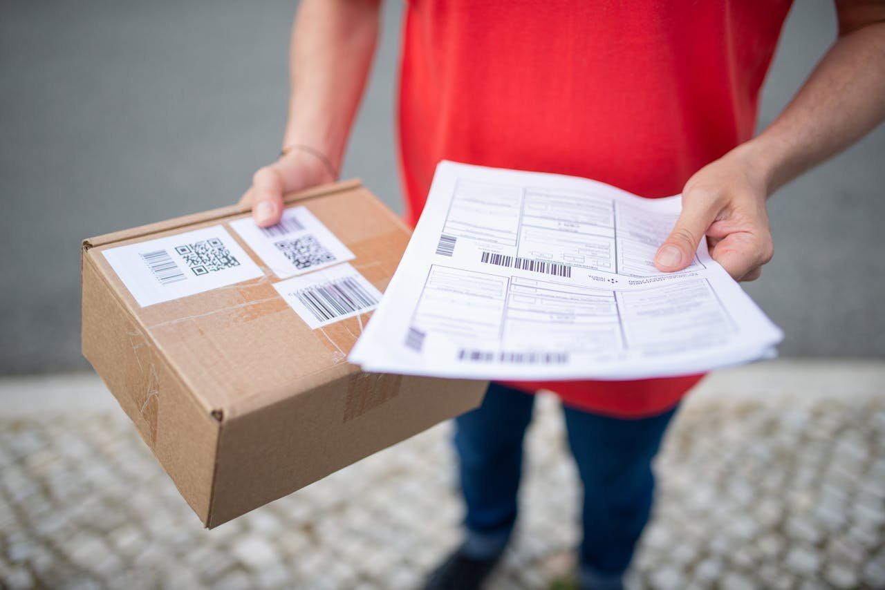Close-up of person in red shirt holding a brown package and delivery documents outside.